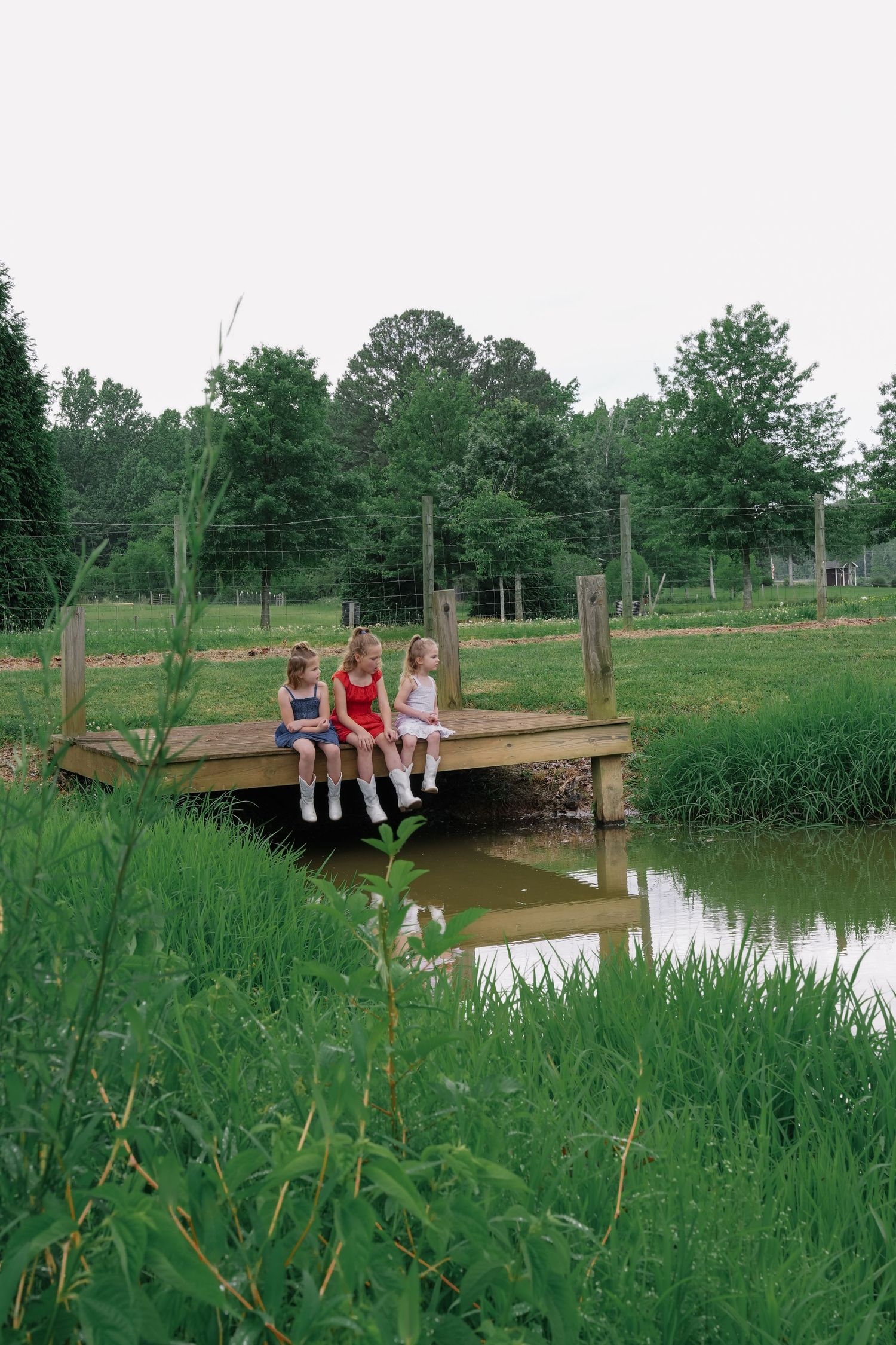 Three people sitting together on a wooden dock overlooking a serene pond surrounded by greenery.