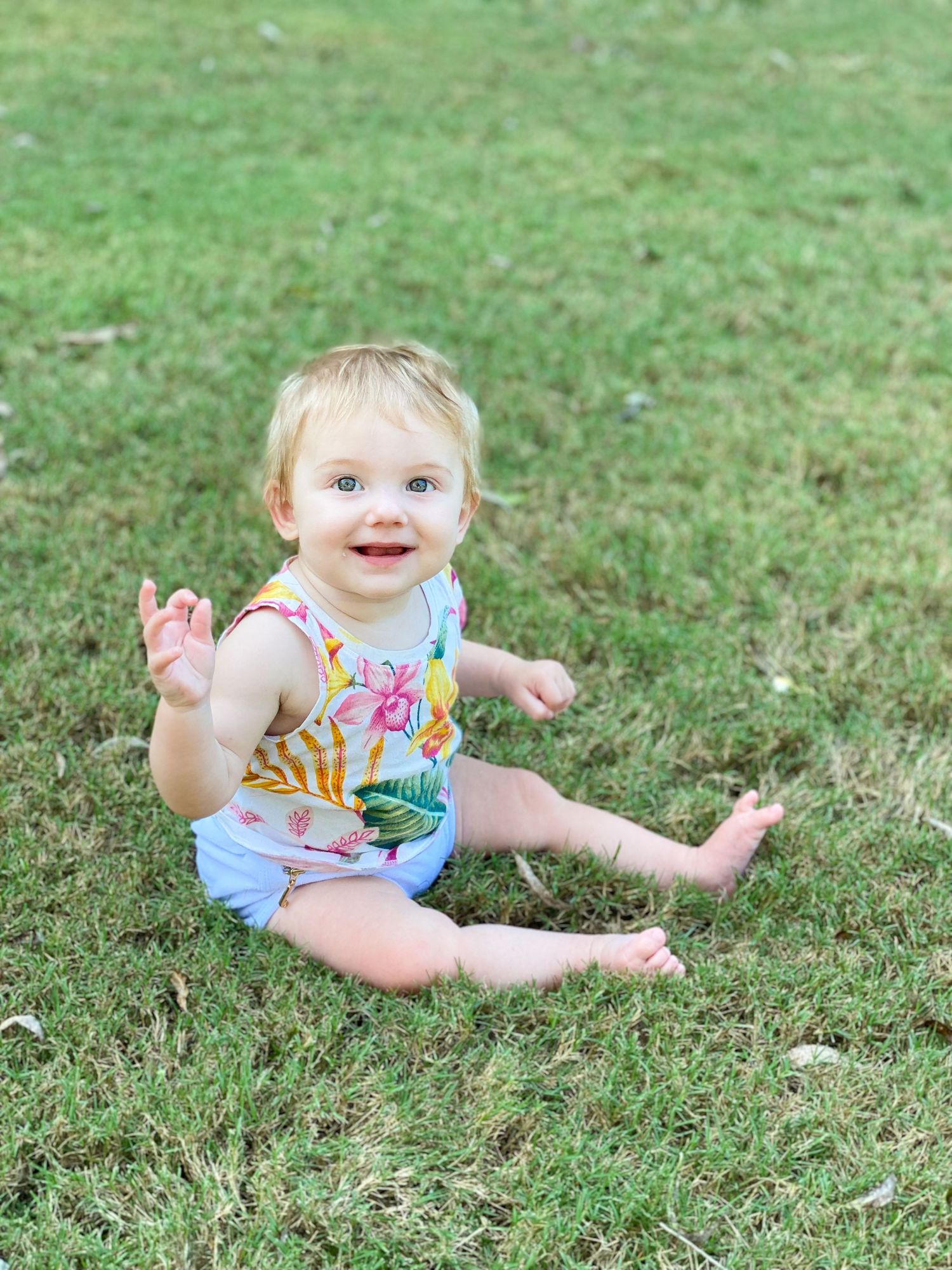 Baby in floral romper sits smiling on green grass on summer day.