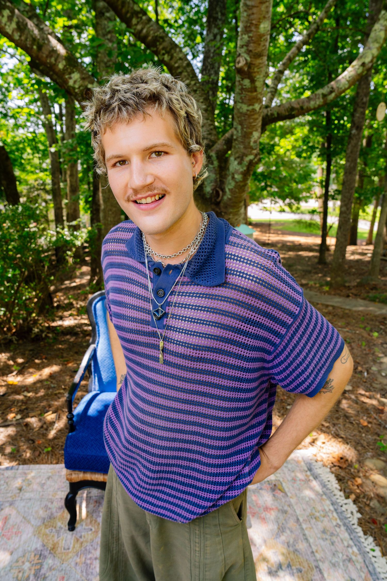 Person wearing striped polo shirt stands in wooded area with sunlit trees in background.