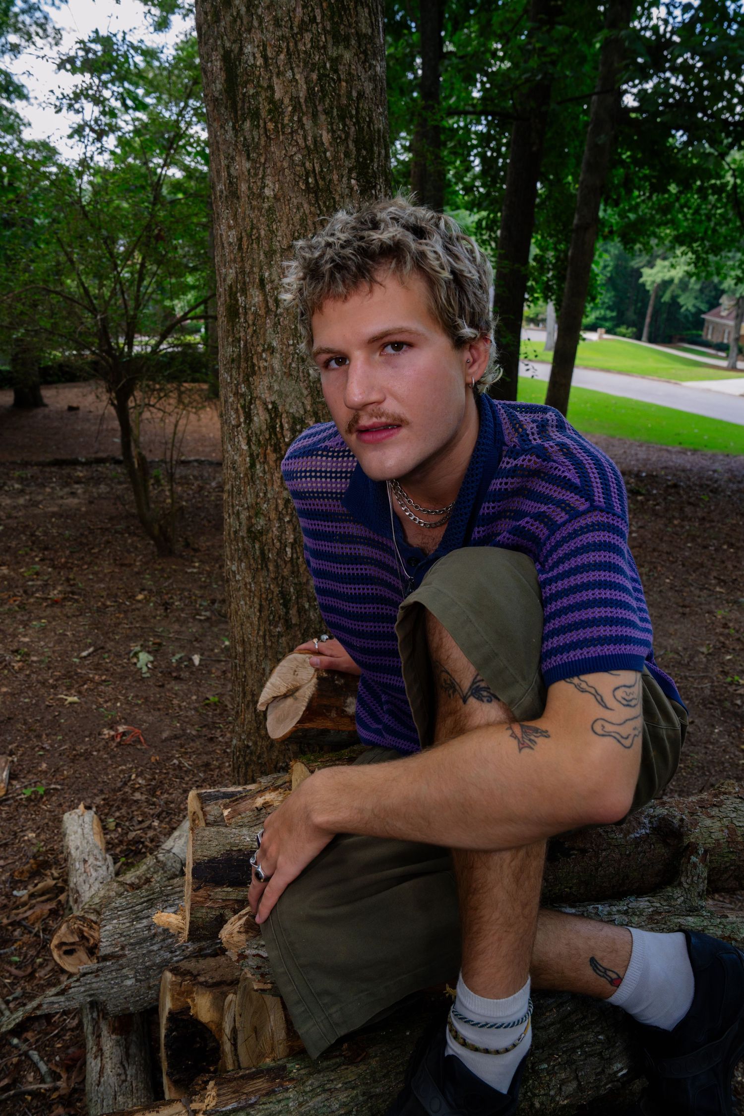 Person in striped purple shirt sits by tree trunk in wooded outdoor setting.