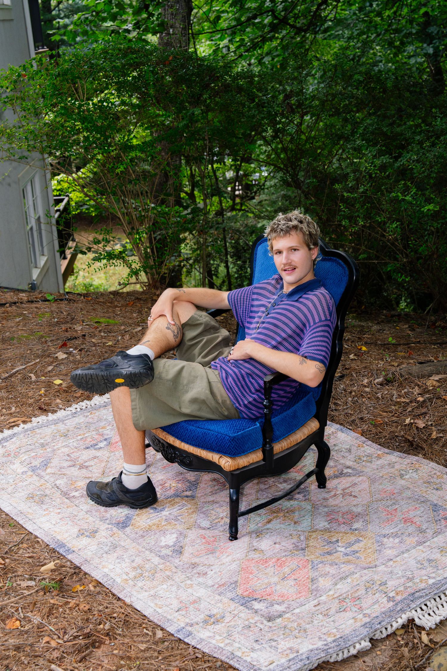 Person lounging in blue chair on patterned rug in a serene garden setting.
