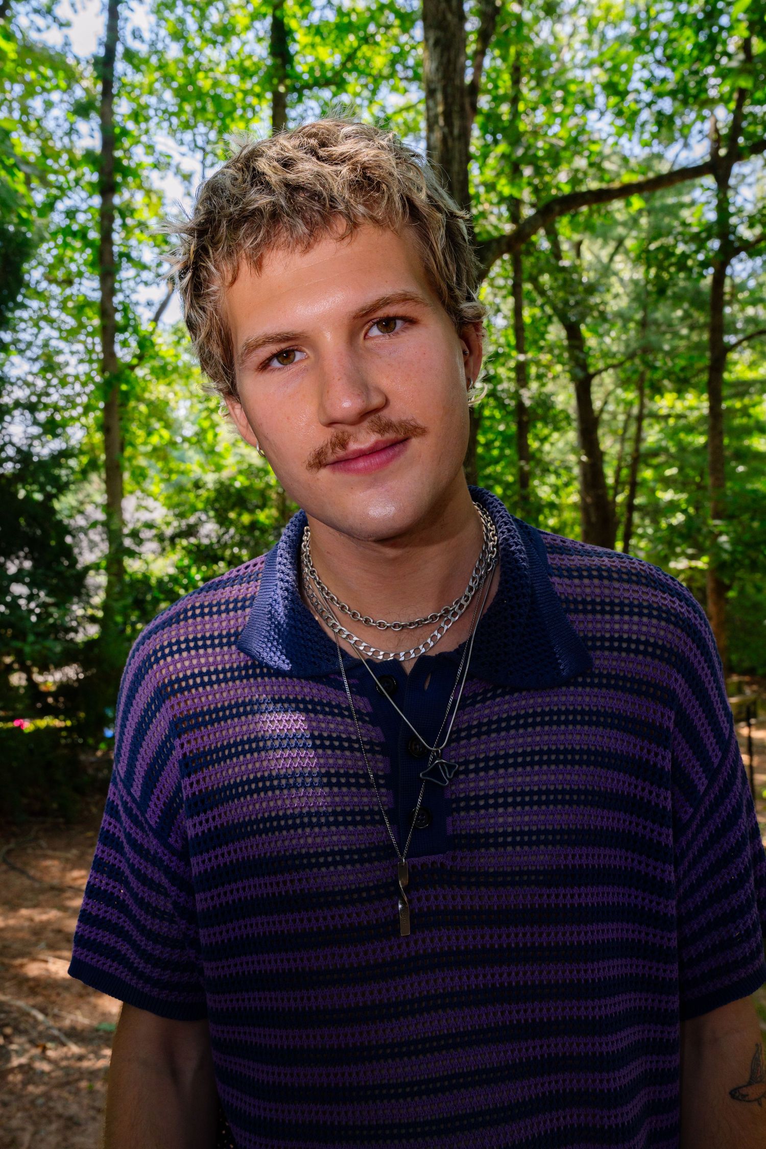Someone in a navy striped shirt poses casually outdoors surrounded by trees.