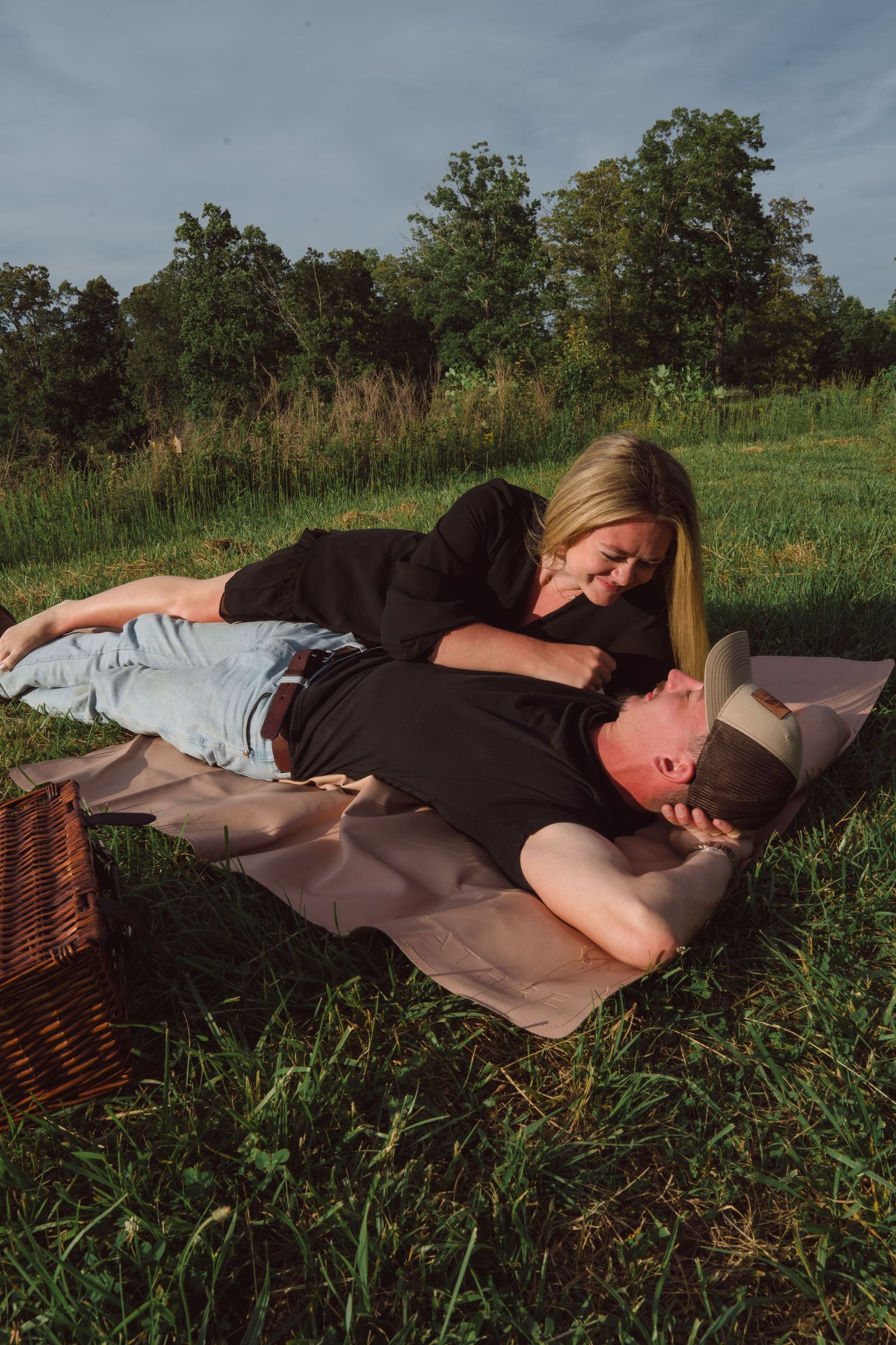 Couple having romantic picnic on blanket with wicker basket in grassy field at dusk.