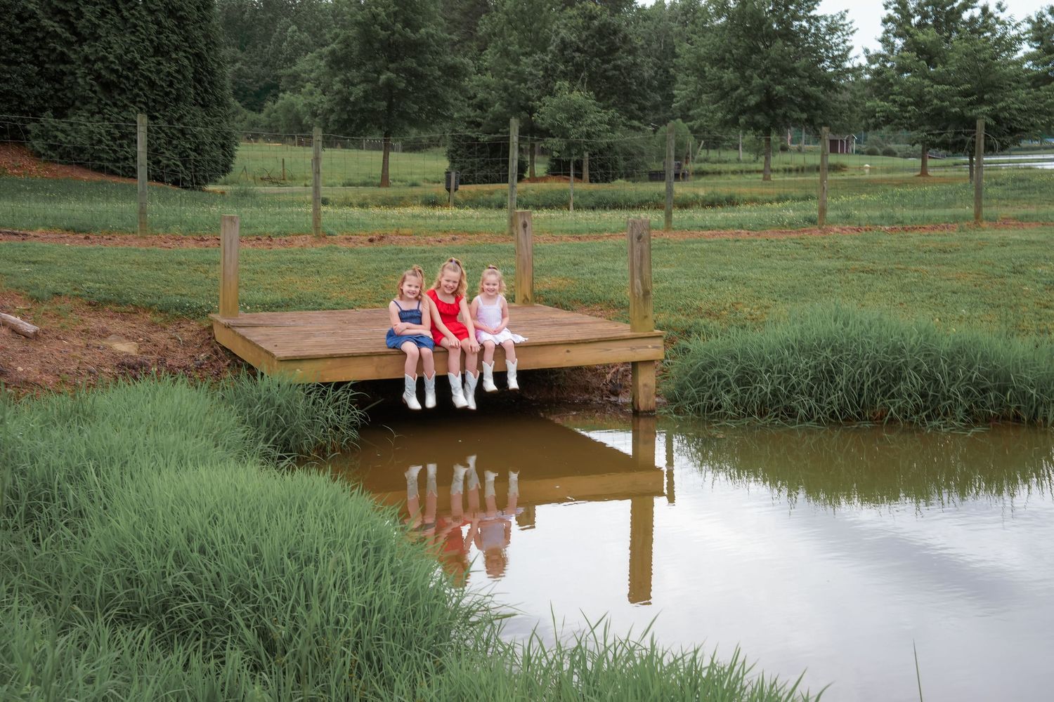 Three figures sitting on wooden dock reflecting in still pond water with green grass and trees.