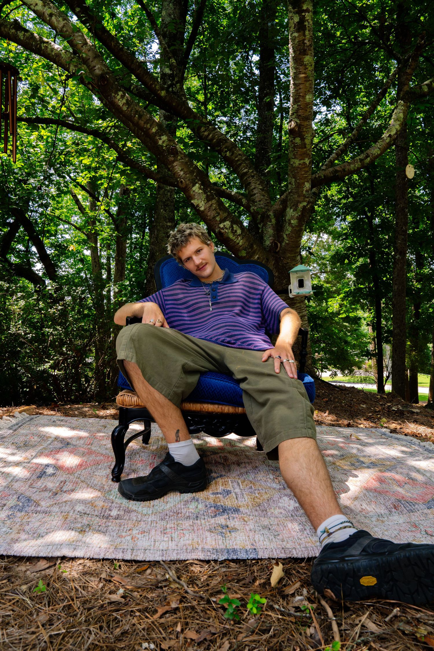 Person relaxing on a patterned rug outdoors while wearing olive pants and a striped polo.