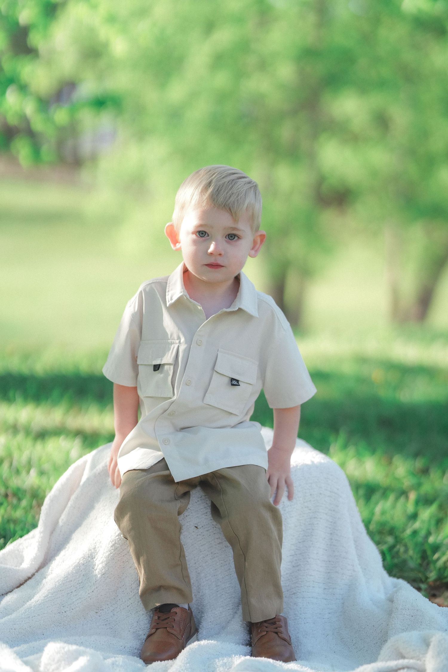 Young child in khaki pants sits on white blanket during outdoor photo session.