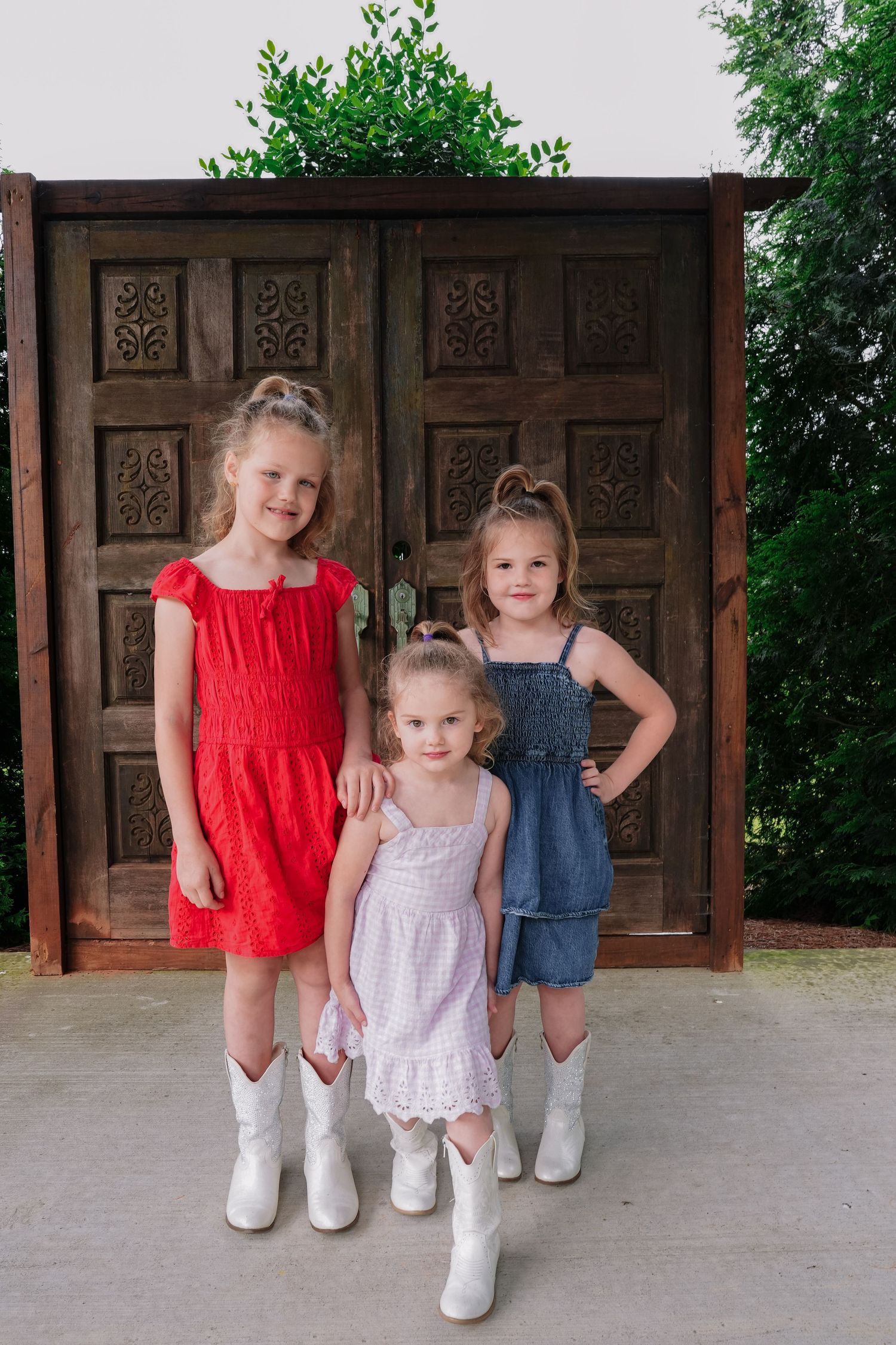 Three children in summer dresses and white boots pose together in front of a wooden carved door.