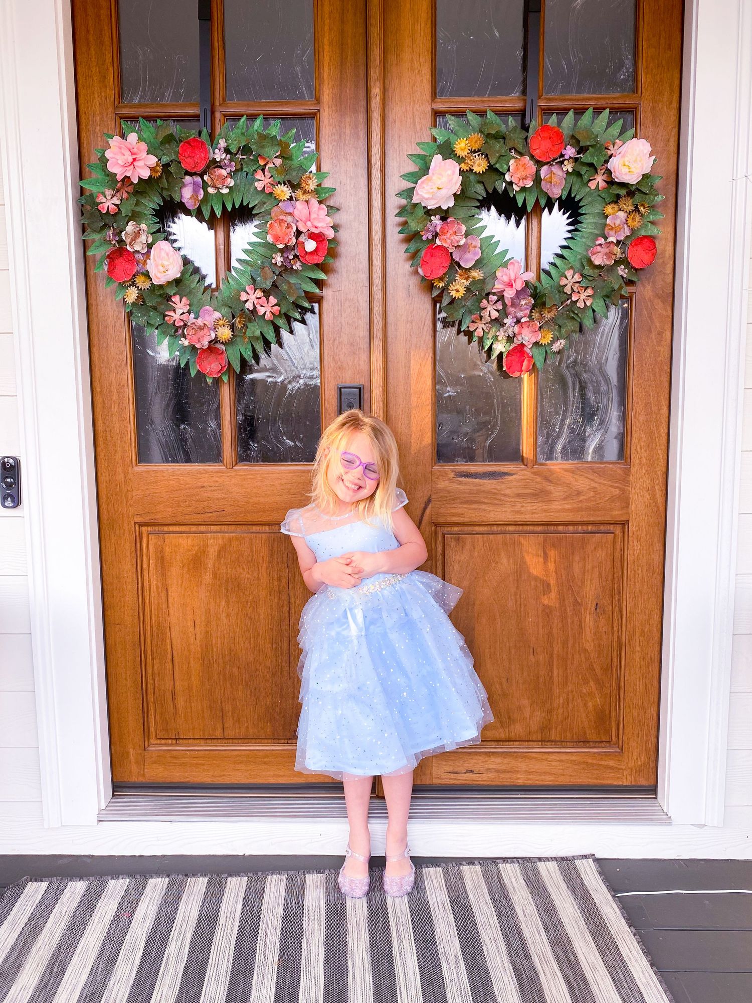 Child in blue tulle dress stands in front of wooden door with floral wreaths.