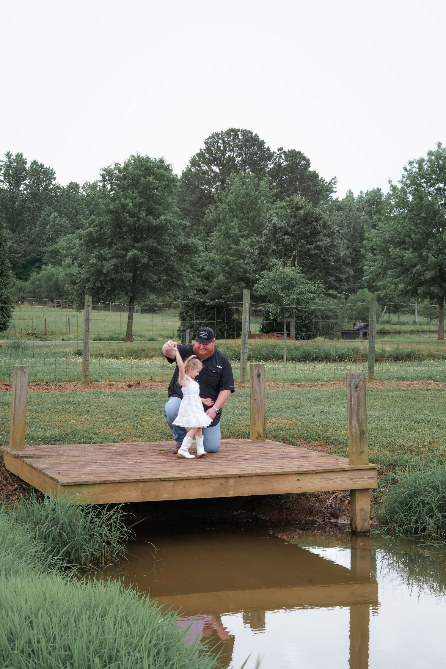 Two people embracing on wooden dock over pond with green pasture and trees in background.