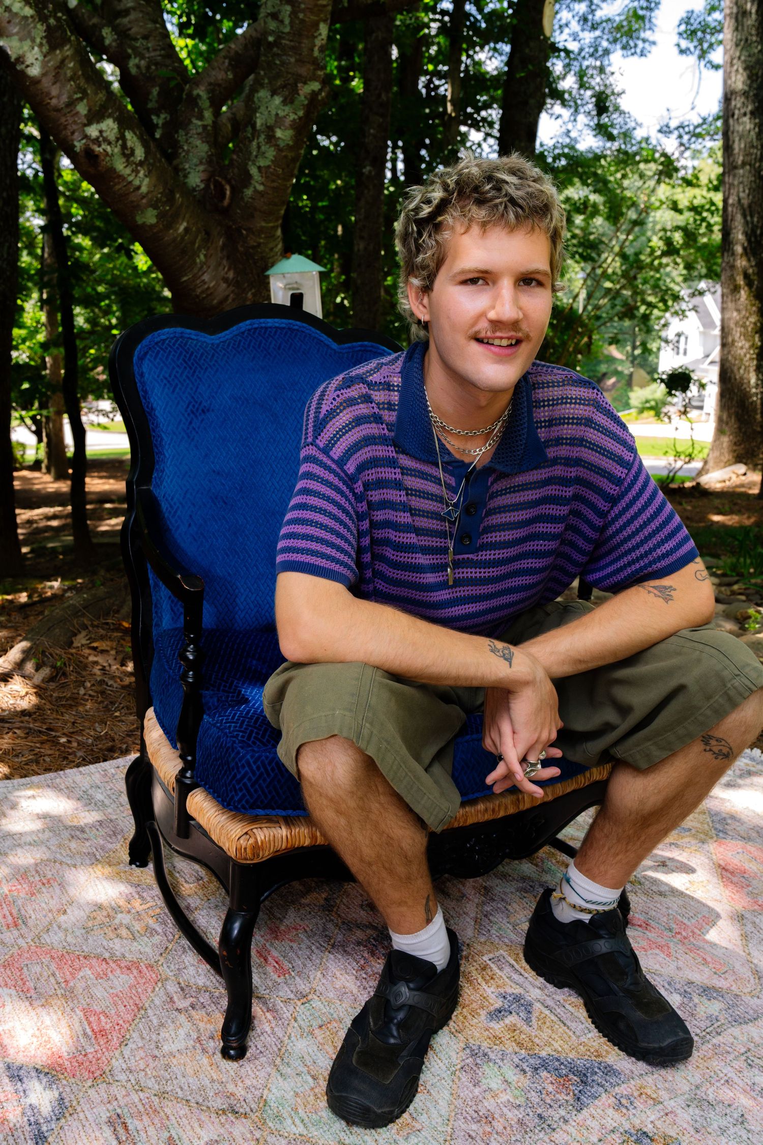 Casual portrait of someone sitting in blue chair on decorative outdoor rug surrounded by trees.