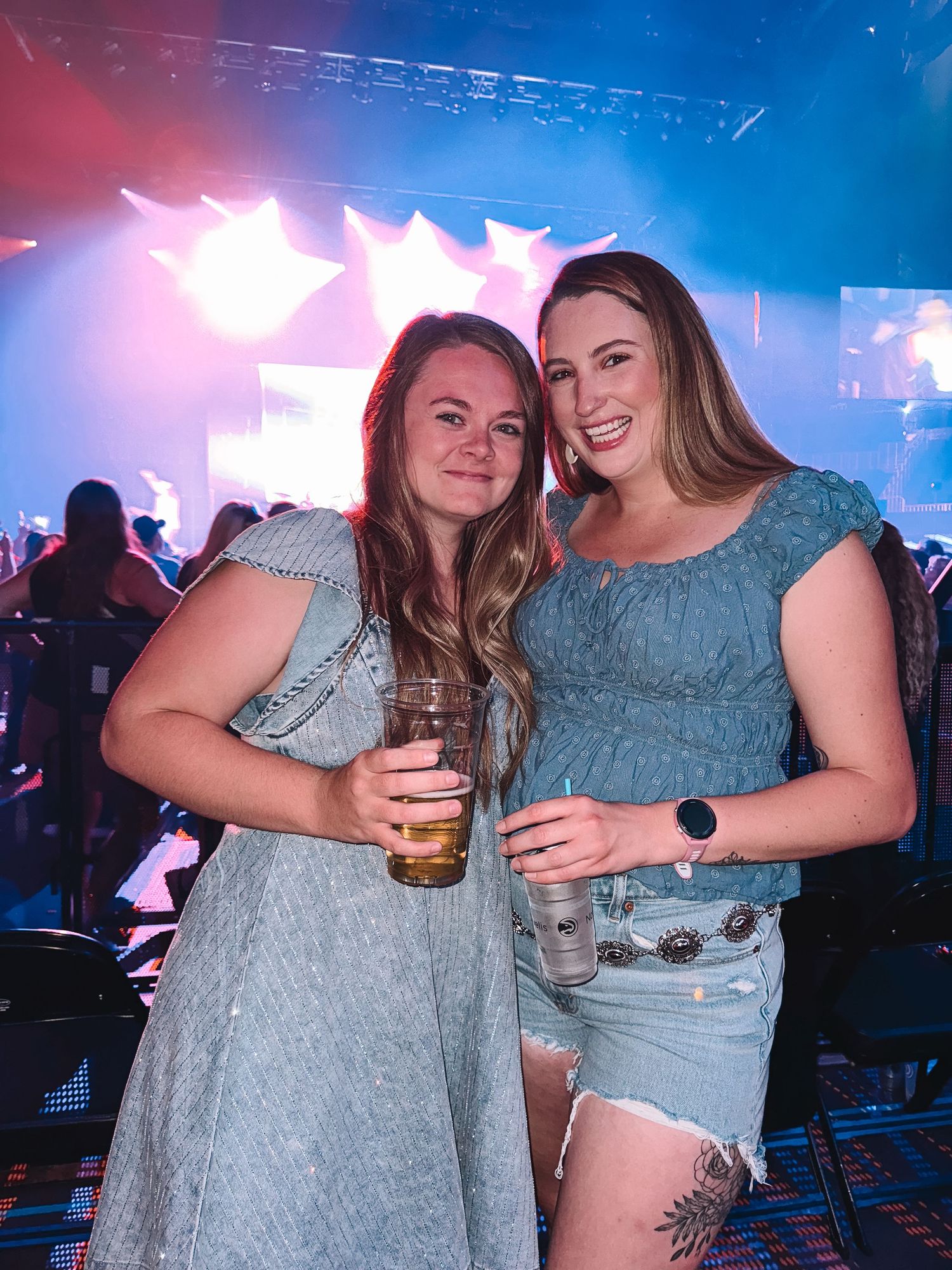 Two friends enjoying drinks at a concert venue with blue and red stage lighting.
