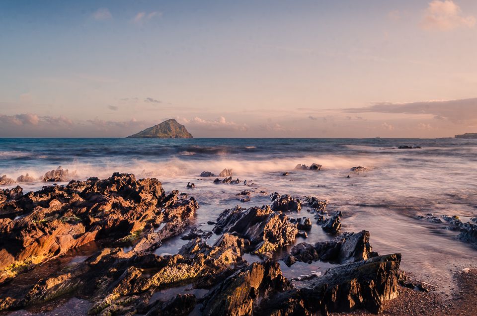 Dramatic panoramic sunset view of rugged coastal cliffs with waves crashing against rocks and misty ocean horizon.