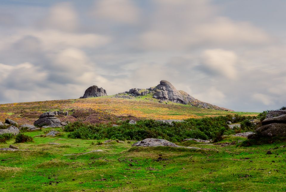 Rocky outcrop rises from green hillside with scattered boulders under cloudy skies in rugged countryside landscape.