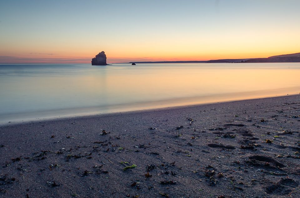 Serene beach at sunset with silhouetted rock formation in distance and smooth water reflecting orange sky.