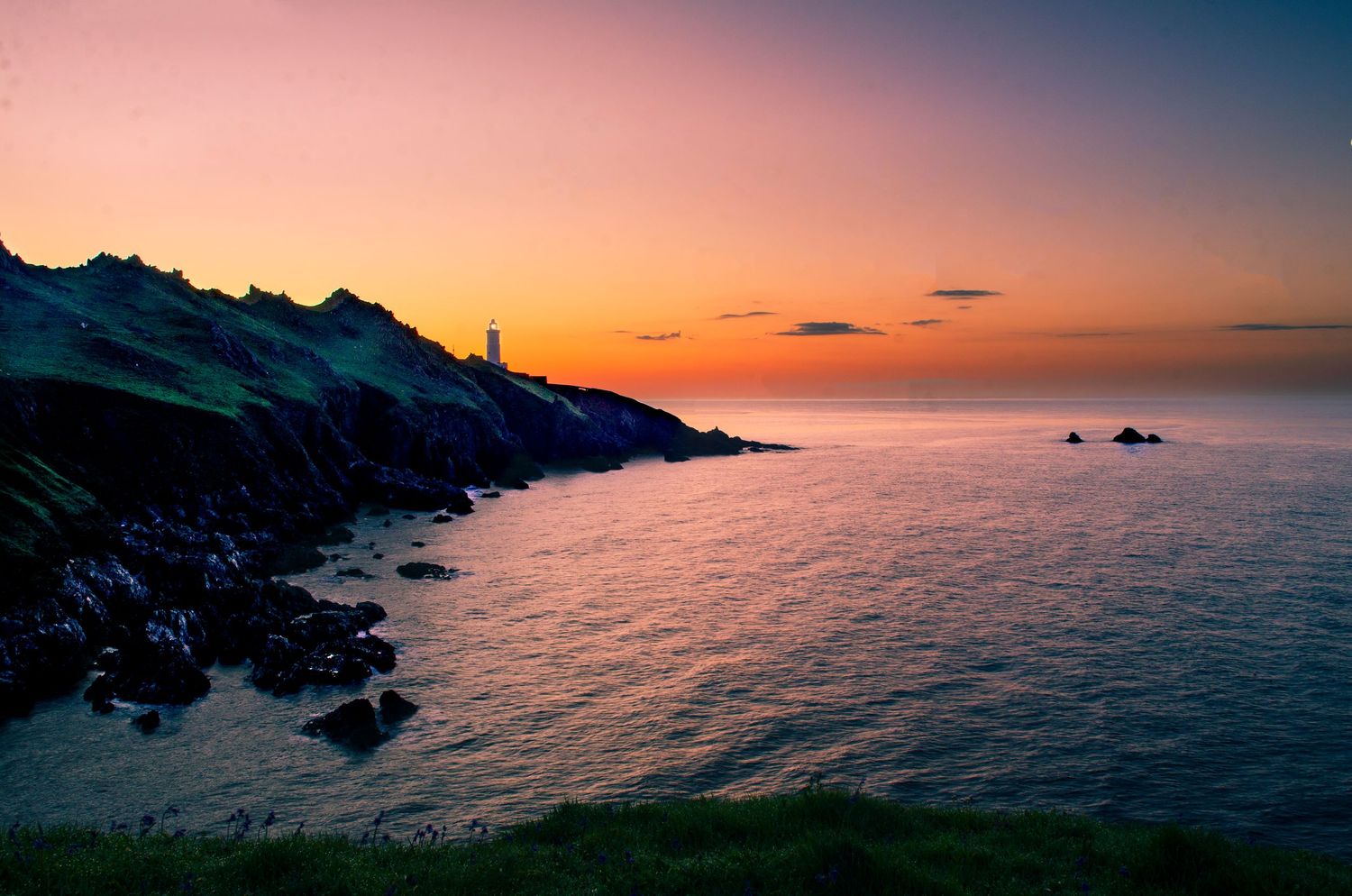 Dramatic sunset over a rocky coastal landscape with lighthouse silhouette and calm ocean waters reflecting orange skies.