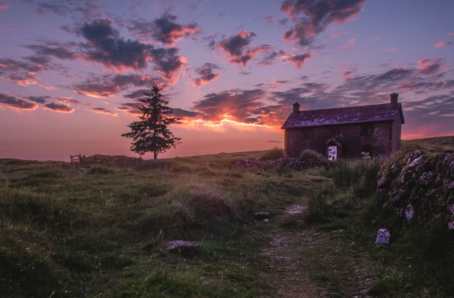 A small rustic cottage on a hilltop at sunset with pink clouds and a lone tree silhouetted against the colorful sky.