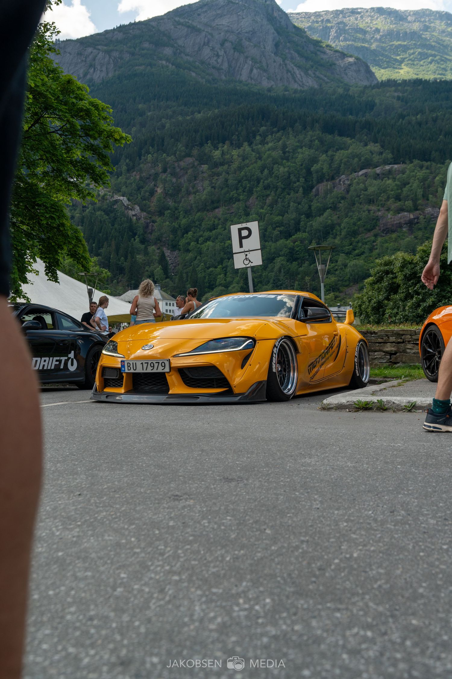 Luxury supercars parked in a mountain parking area with dramatic mountain scenery in the background.