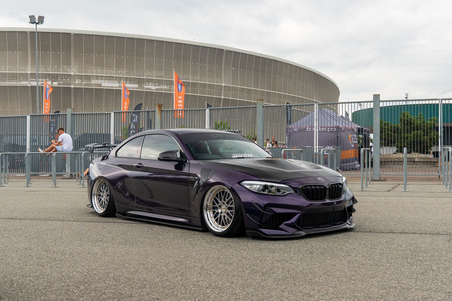 Two stanced BMW cars, one in deep purple and one in black, displayed on a lot with custom wheels.
