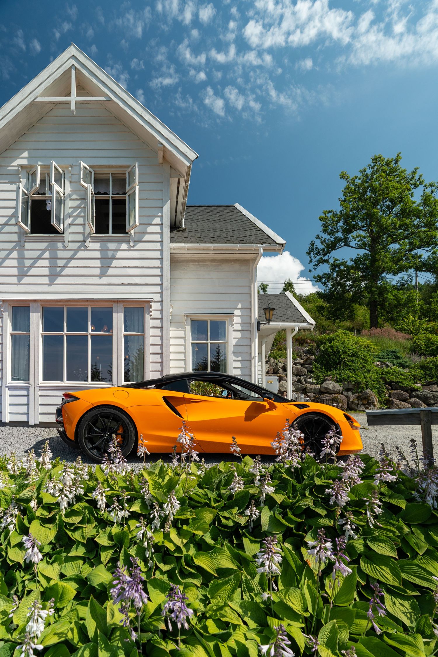 Orange McLaren sports car parked beside a white wooden house with lush green gardens and purple flowers under a blue sky.