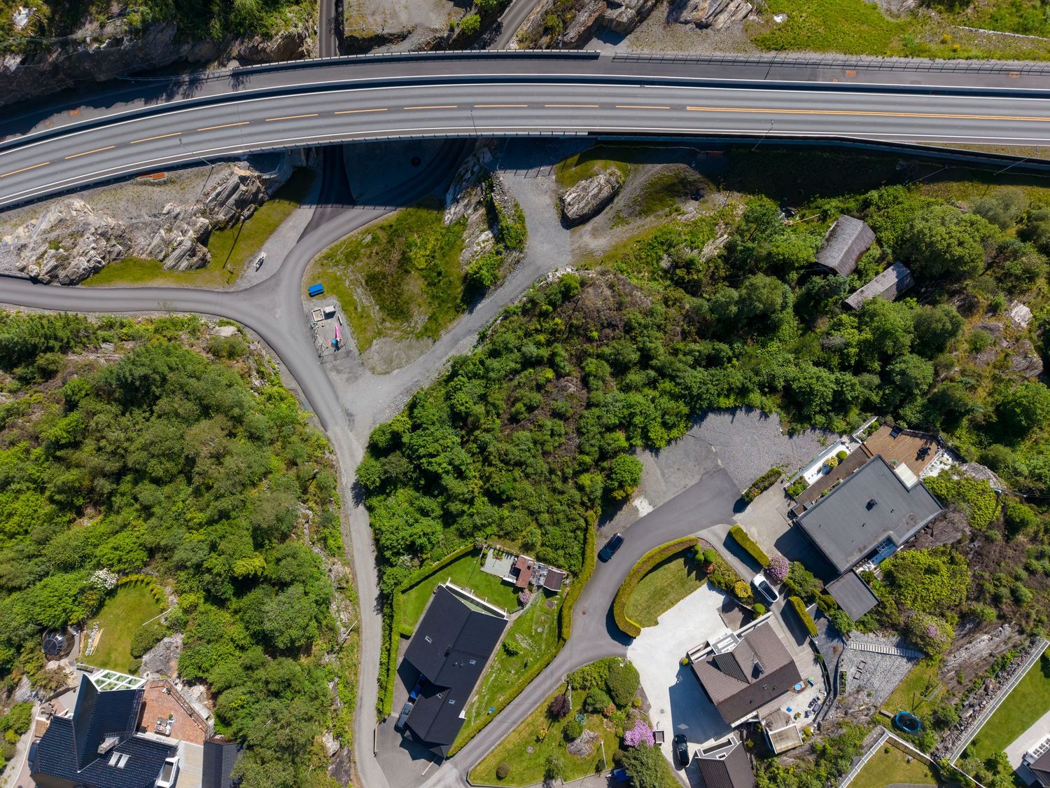Aerial view of a highway bridge curving above a green hillside with scattered houses, winding roads, and dense trees in a coastal village.