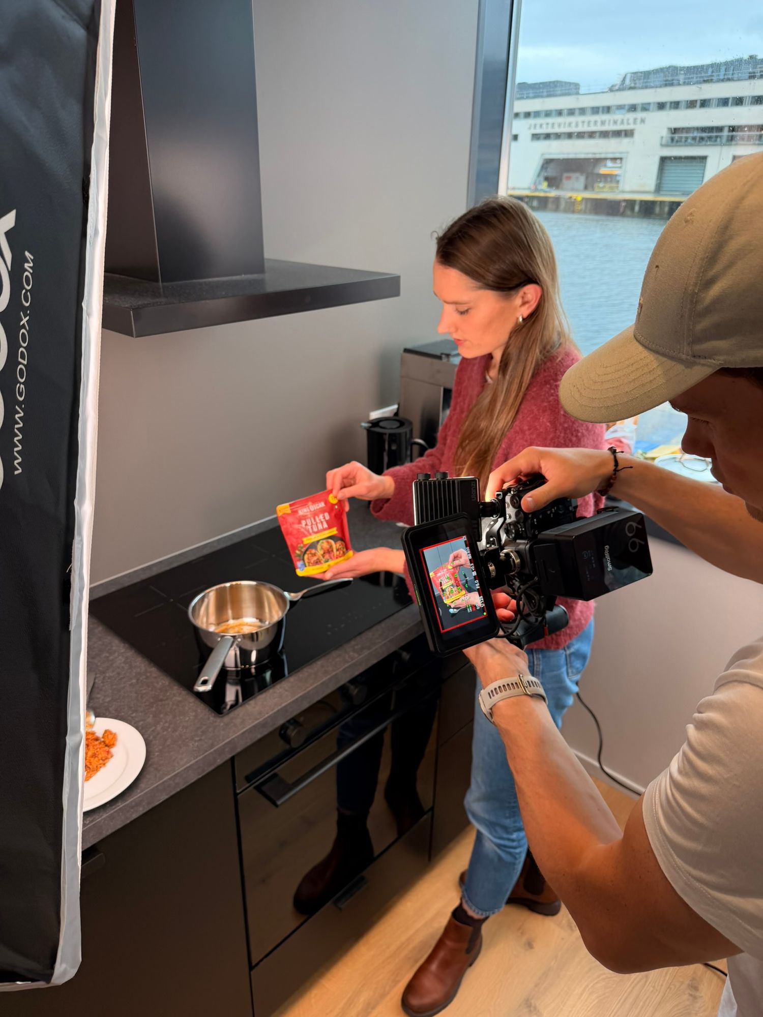 Woman holding a red food package being filmed by a cameraman in a modern kitchen studio near a waterfront.