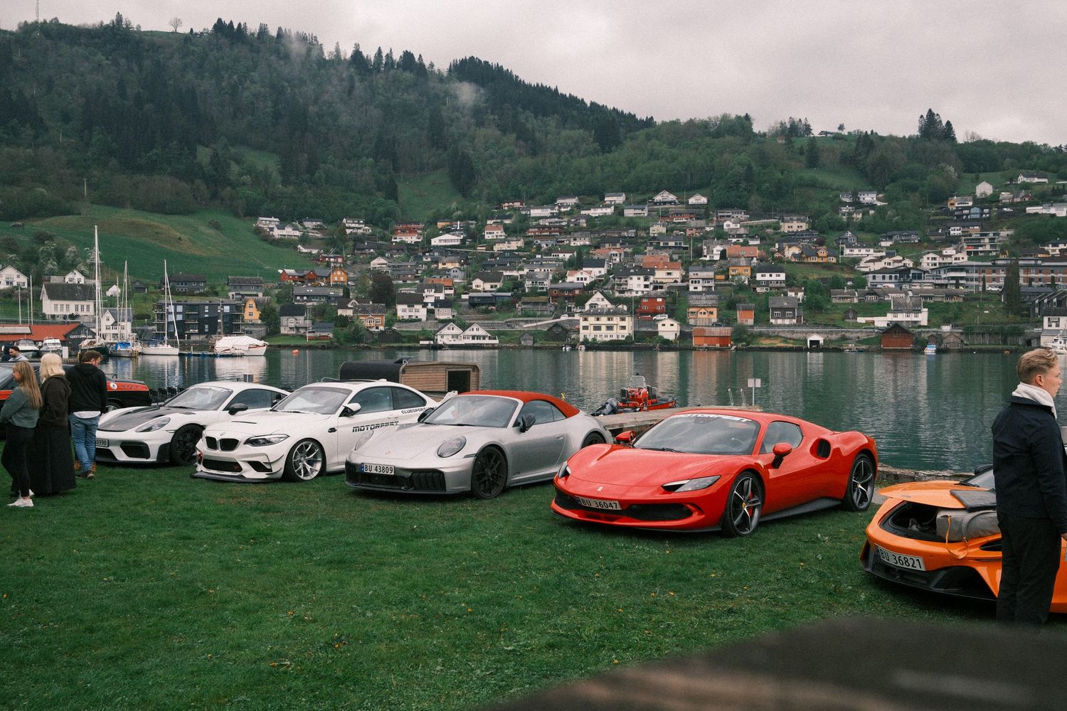 Red, silver and orange sports cars parked on a lakeside lawn with a hillside village, boats and forested mountains in the background.