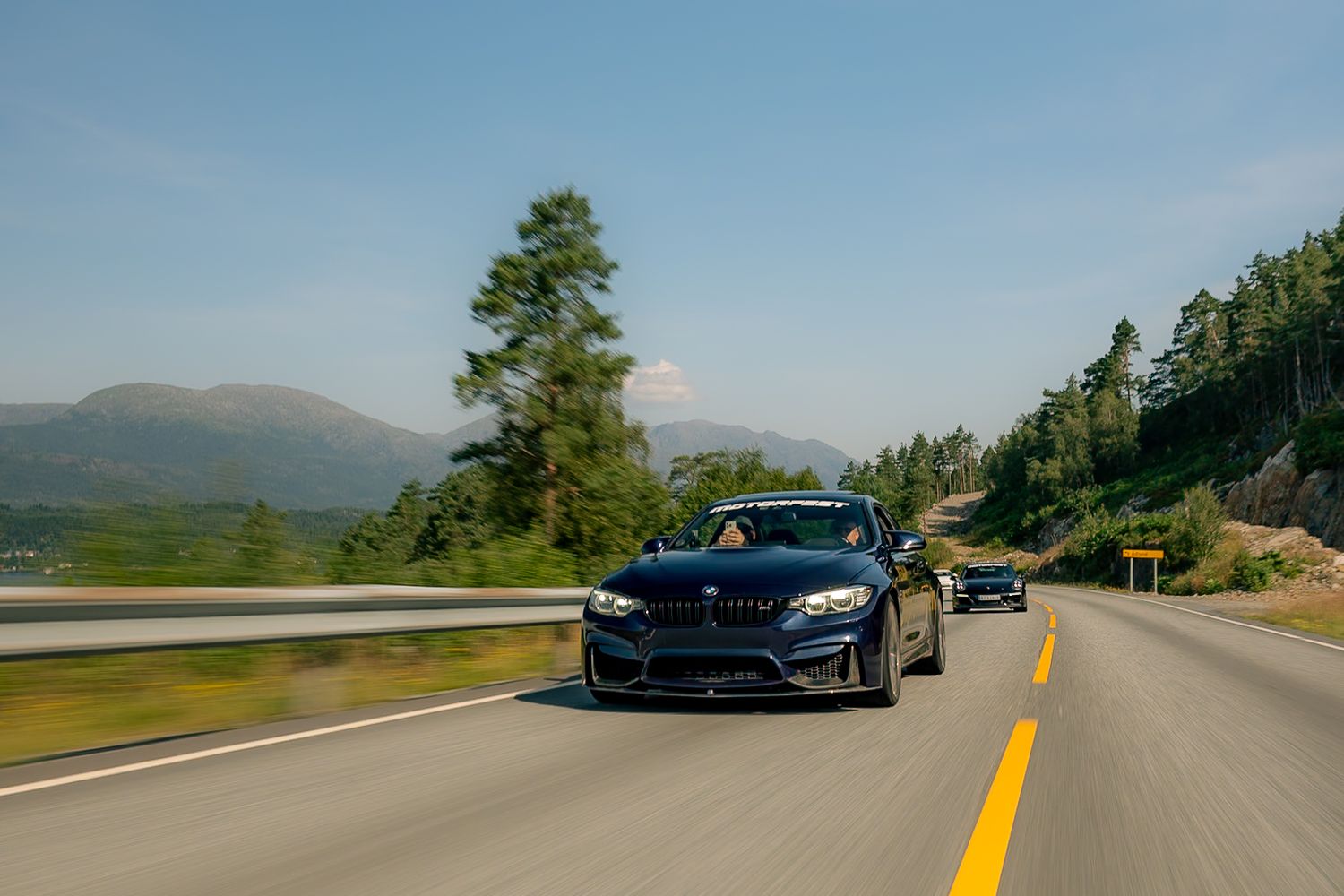 Dark blue BMW sports sedan driving along a curving mountain road with forested hills and distant peaks under a clear blue sky.