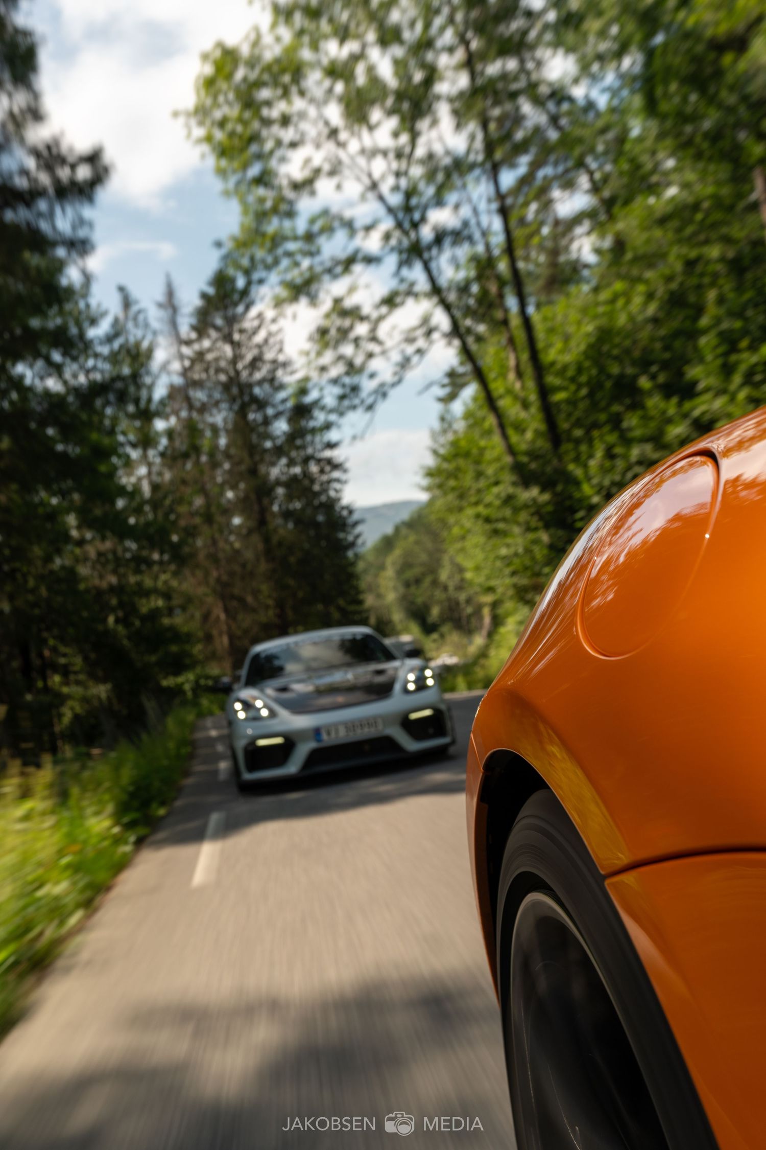 POV driving shot showing exotic cars on a tree-lined mountain road with motion blur effects.