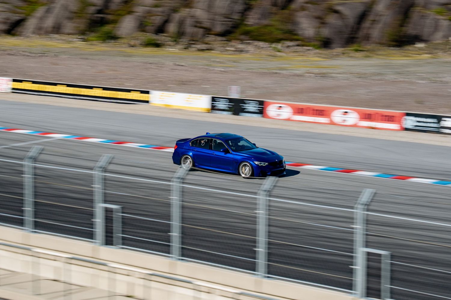 A bright blue sports car races around a track with safety barriers and red and white curbing visible.