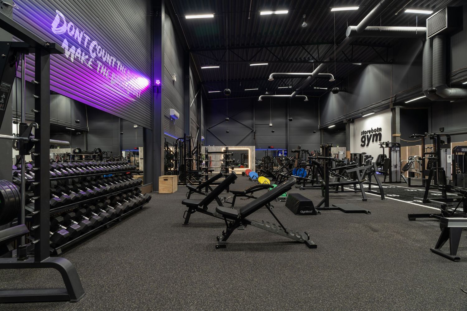 Modern fitness gym interior with purple signage, weight racks, and exercise equipment on dark carpet flooring.