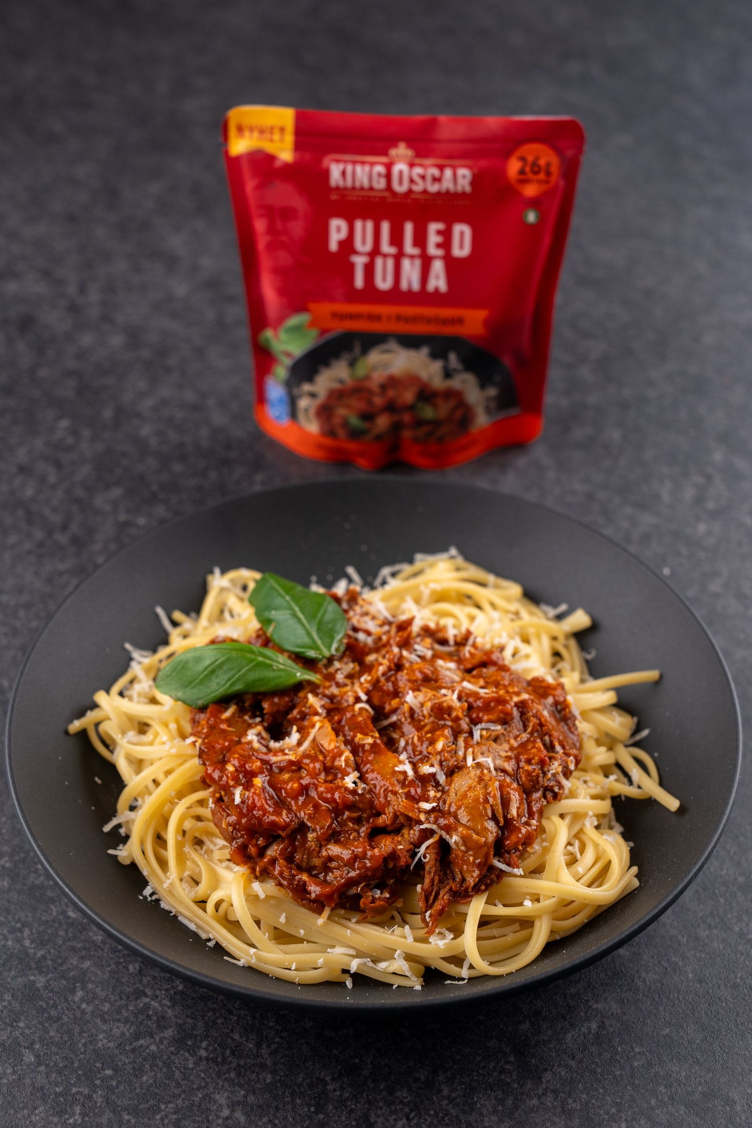 Spaghetti topped with King Oscar pulled tuna in rich tomato sauce, grated cheese and fresh basil, served beside the red pouch.