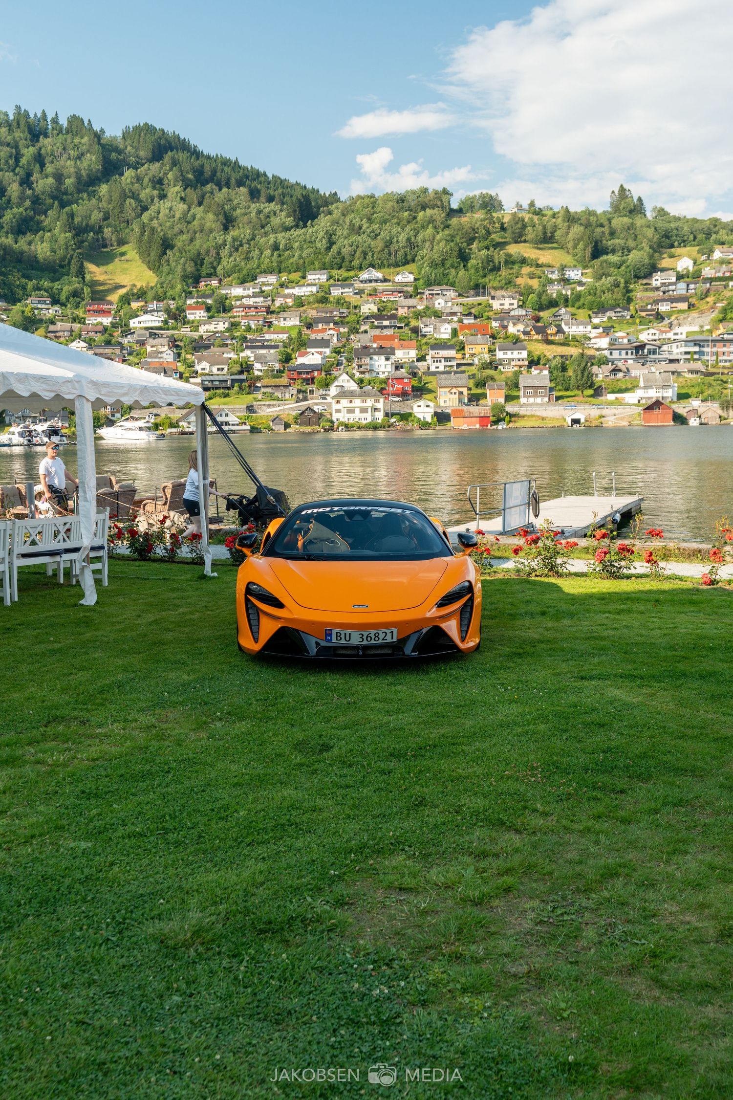 Orange McLaren supercar parked on grass with Norwegian fjord village in background.