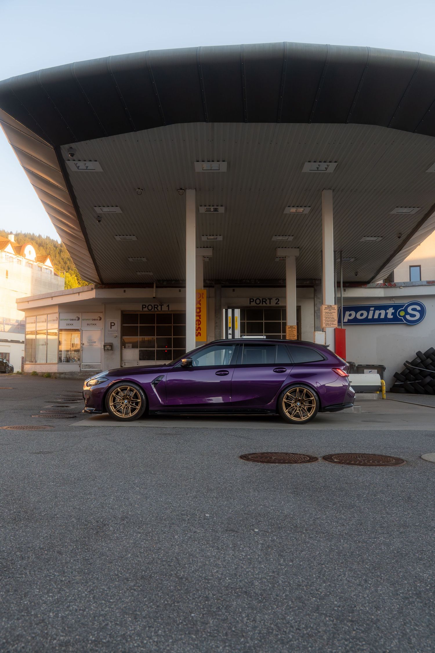 A sleek purple BMW M3 parked under a covered area at sunset with gold wheels and lowered suspension.