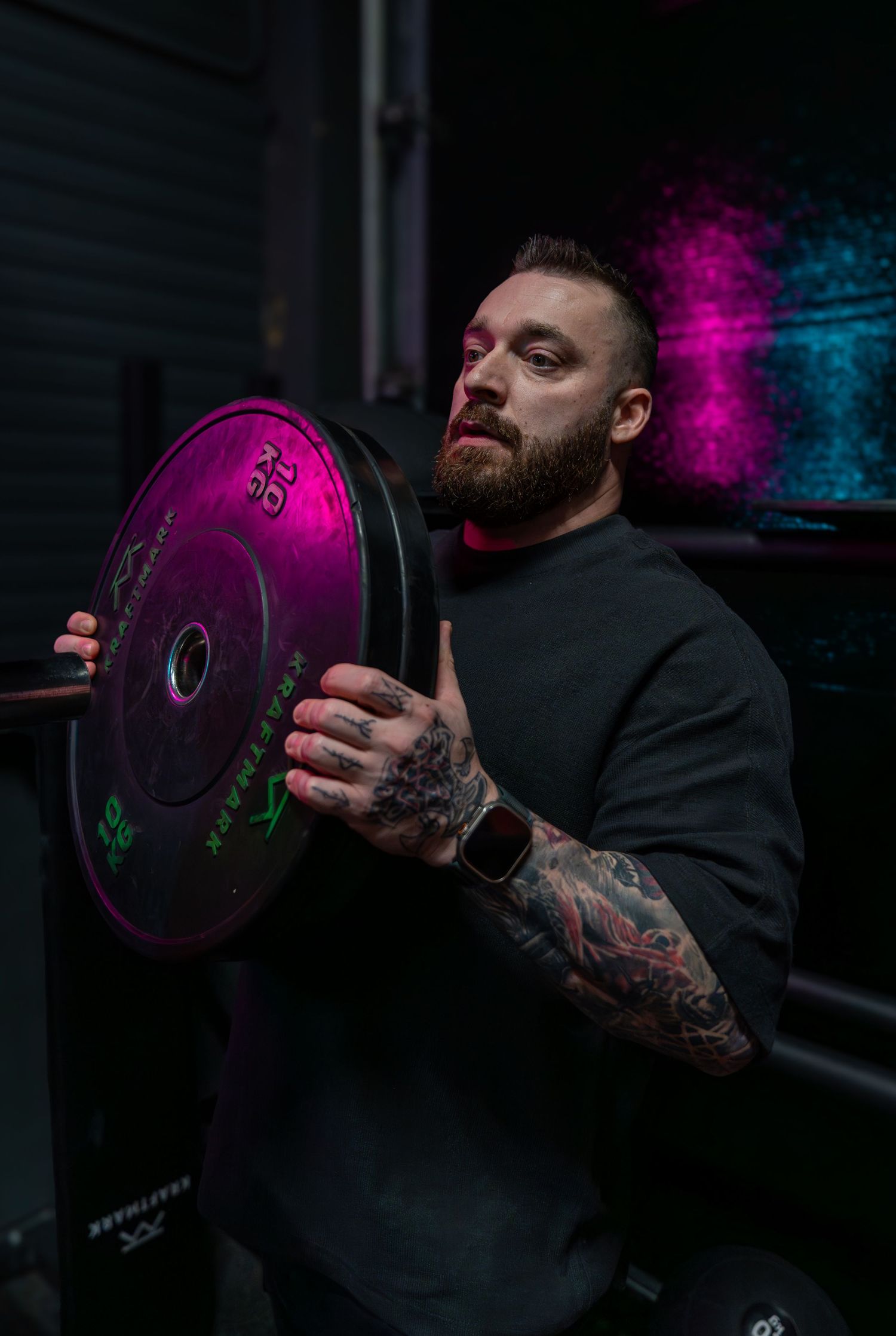 Person holding colored weight plate with detailed sleeve tattoo in dramatic gym lighting.