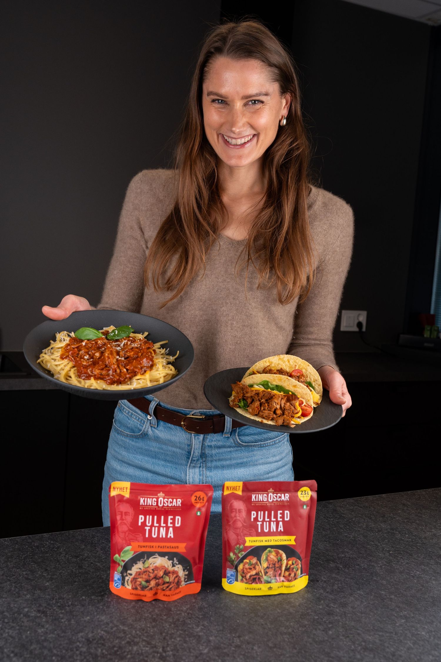 Smiling woman holds a plate of spaghetti with sauce and a pulled tuna taco above two King Oscar Pulled Tuna pouches on a kitchen counter.