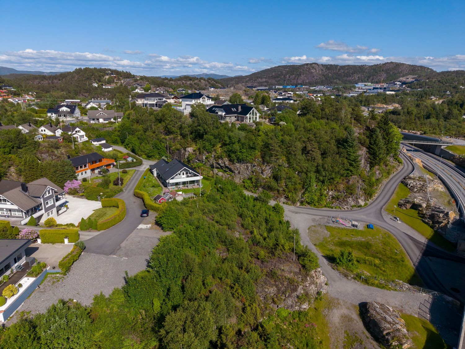 Drone view of a coastal neighborhood with houses on a green rocky hillside, winding roads, and a bridge nearby.