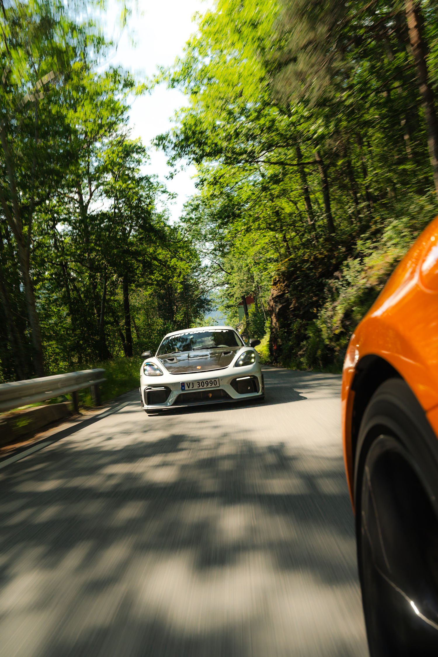 White sports car driving along a tree-lined mountain road in sunlight, with the blurred edge of an orange car in the foreground.