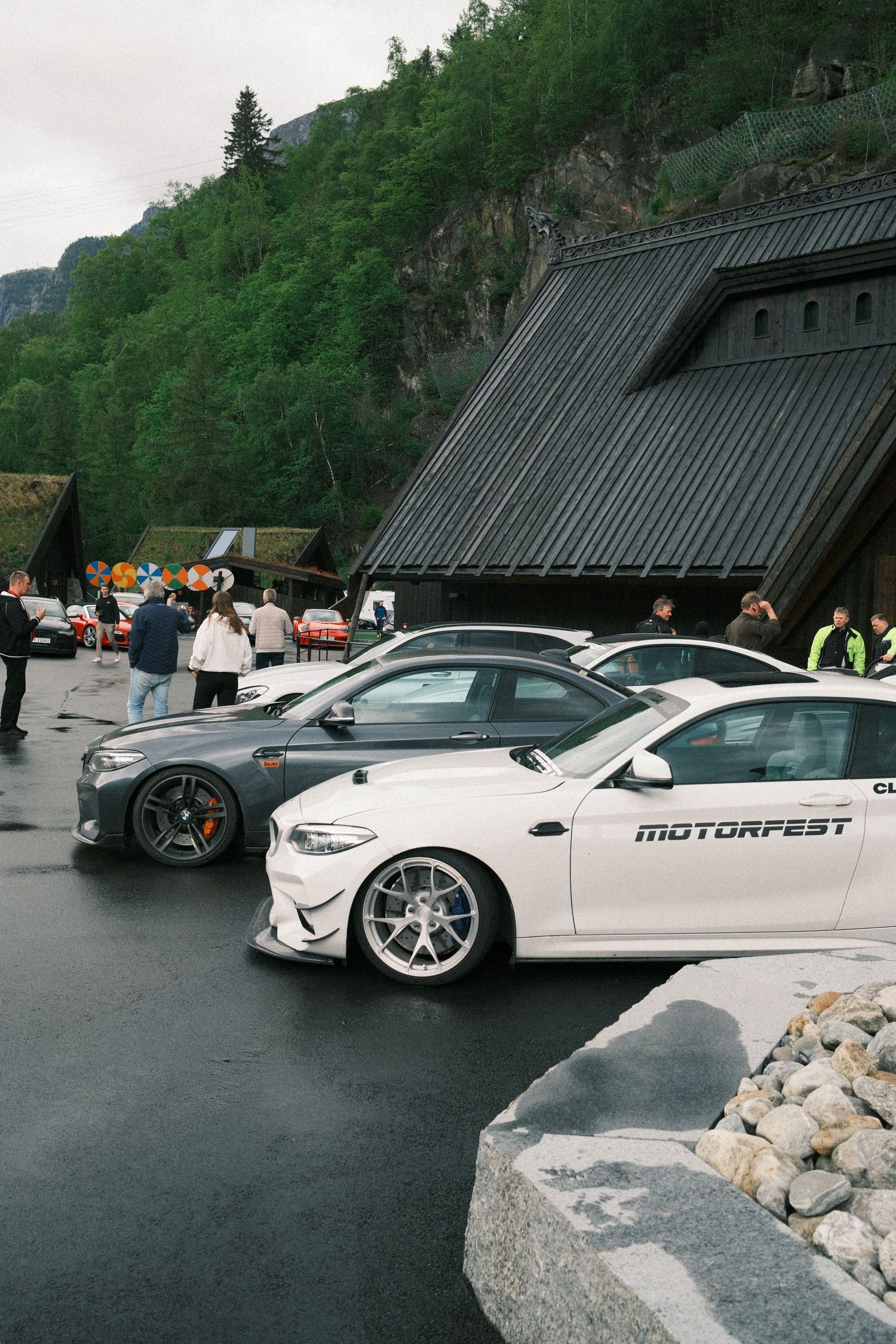 White BMW with Motorfest decals parked beside a gray sports car on wet pavement near a dark wooden building and forested mountain.