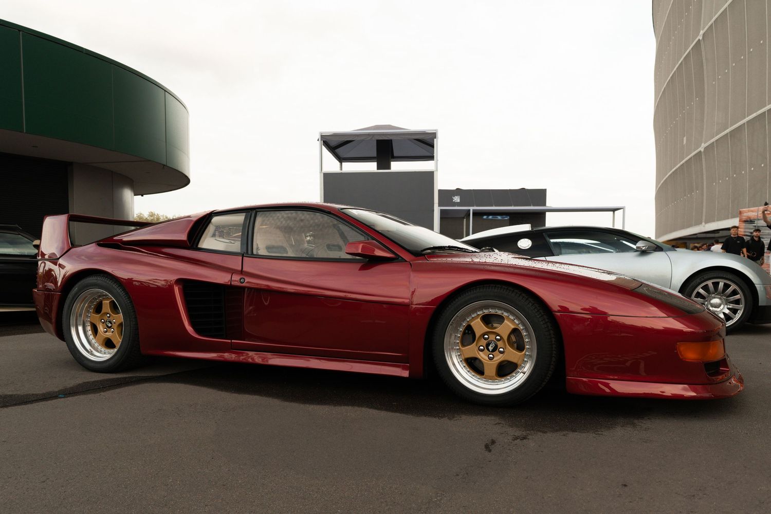 Heavily modified dark red Ferrari Testarossa supercar with widebody kit and gold wheels parked outside a modern building.