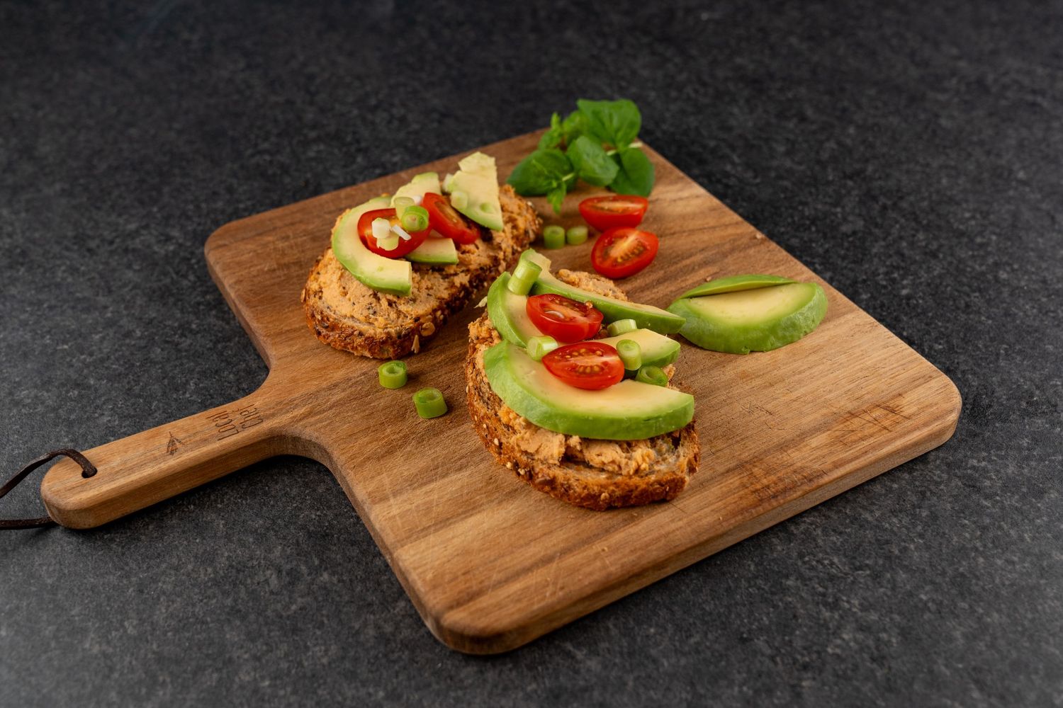 Two avocado toast slices with cherry tomatoes on a wooden cutting board, garnished with fresh herbs and chili peppers.