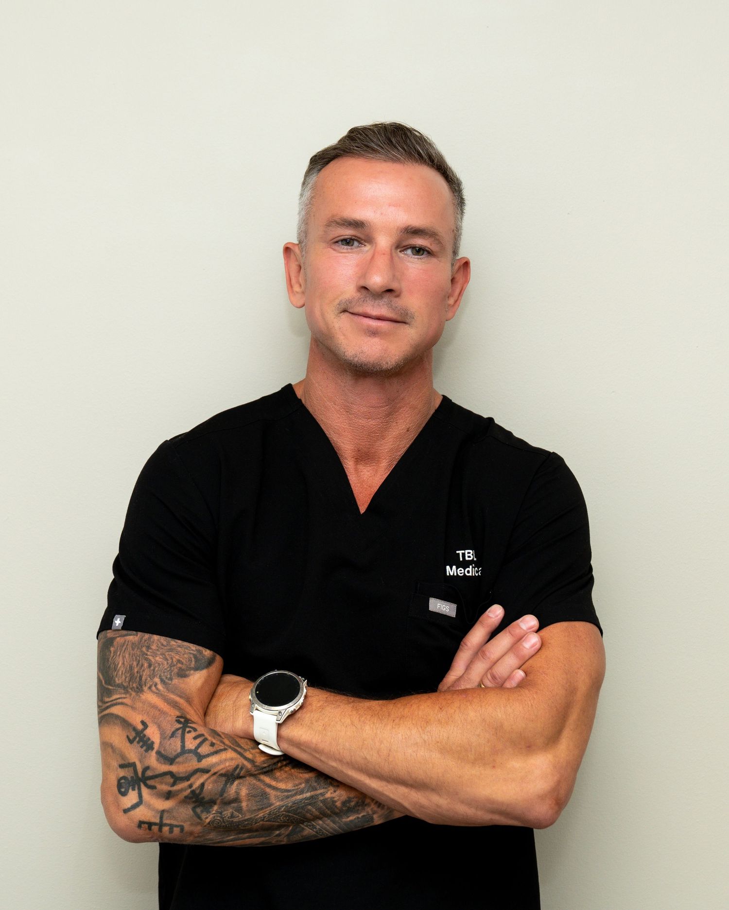 Man in black medical scrubs with crossed arms, tattooed forearm and smartwatch, smiling confidently against a plain light background.