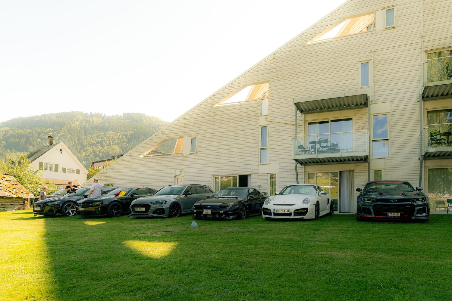 Row of sports cars parked on a sunlit green lawn in front of a modern wooden-clad building with balconies and wooded hills behind.