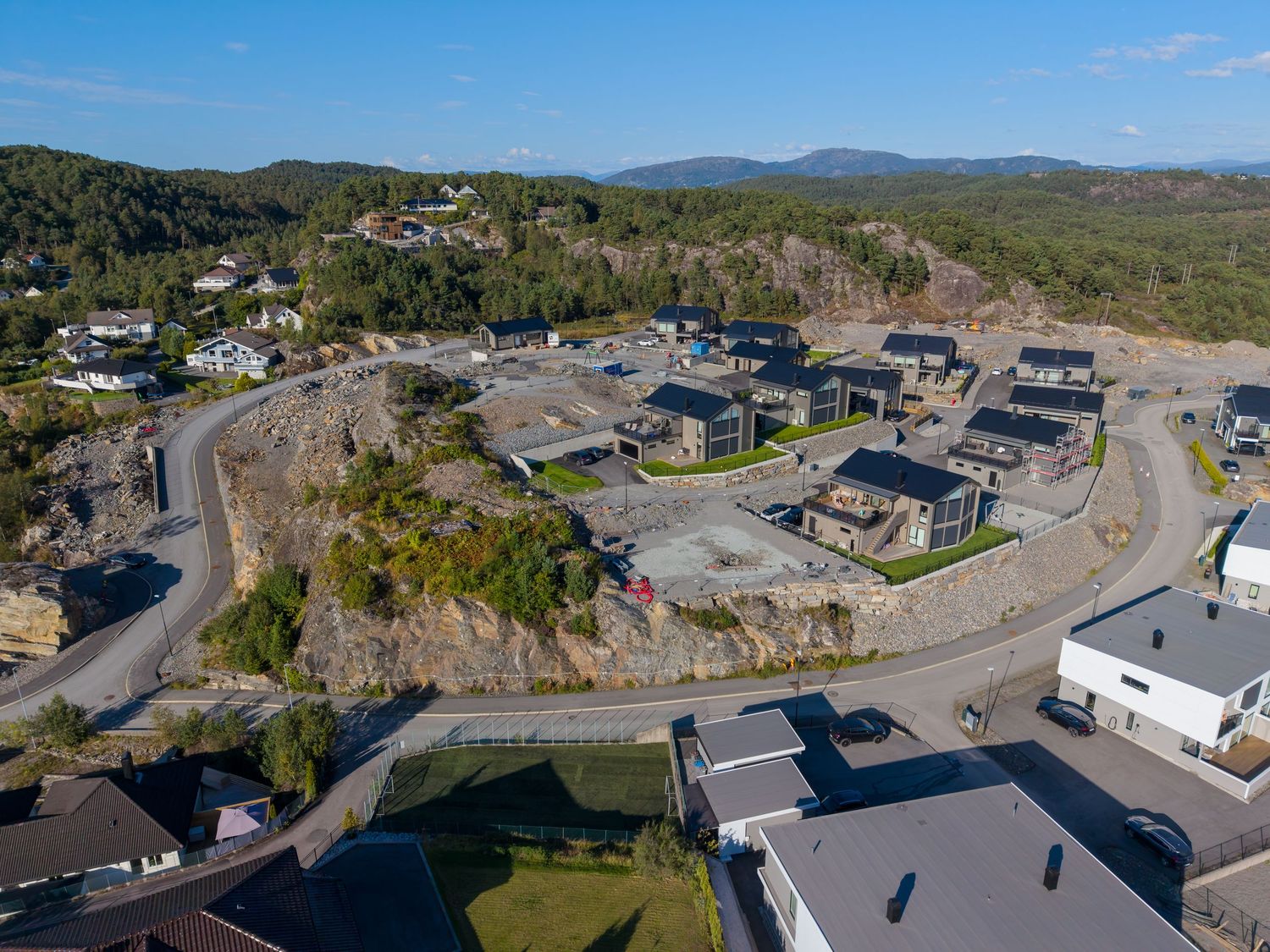 Aerial view of a modern hillside residential neighborhood with new homes, winding roads, and forested hills under a clear blue sky.
