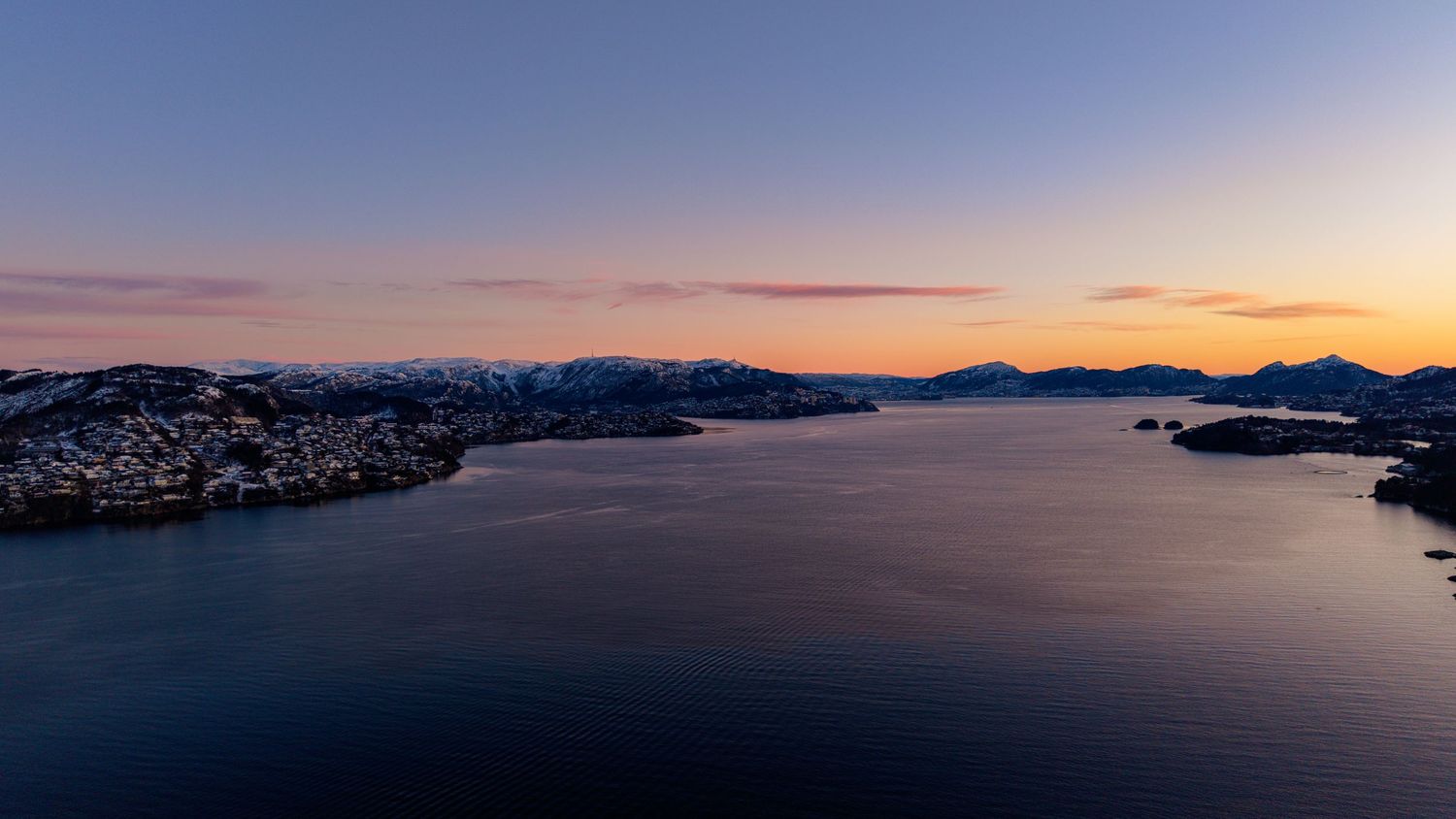 Aerial sunset view over a Norwegian fjord with snow-capped mountains, calm blue waters, and coastal town at dusk.