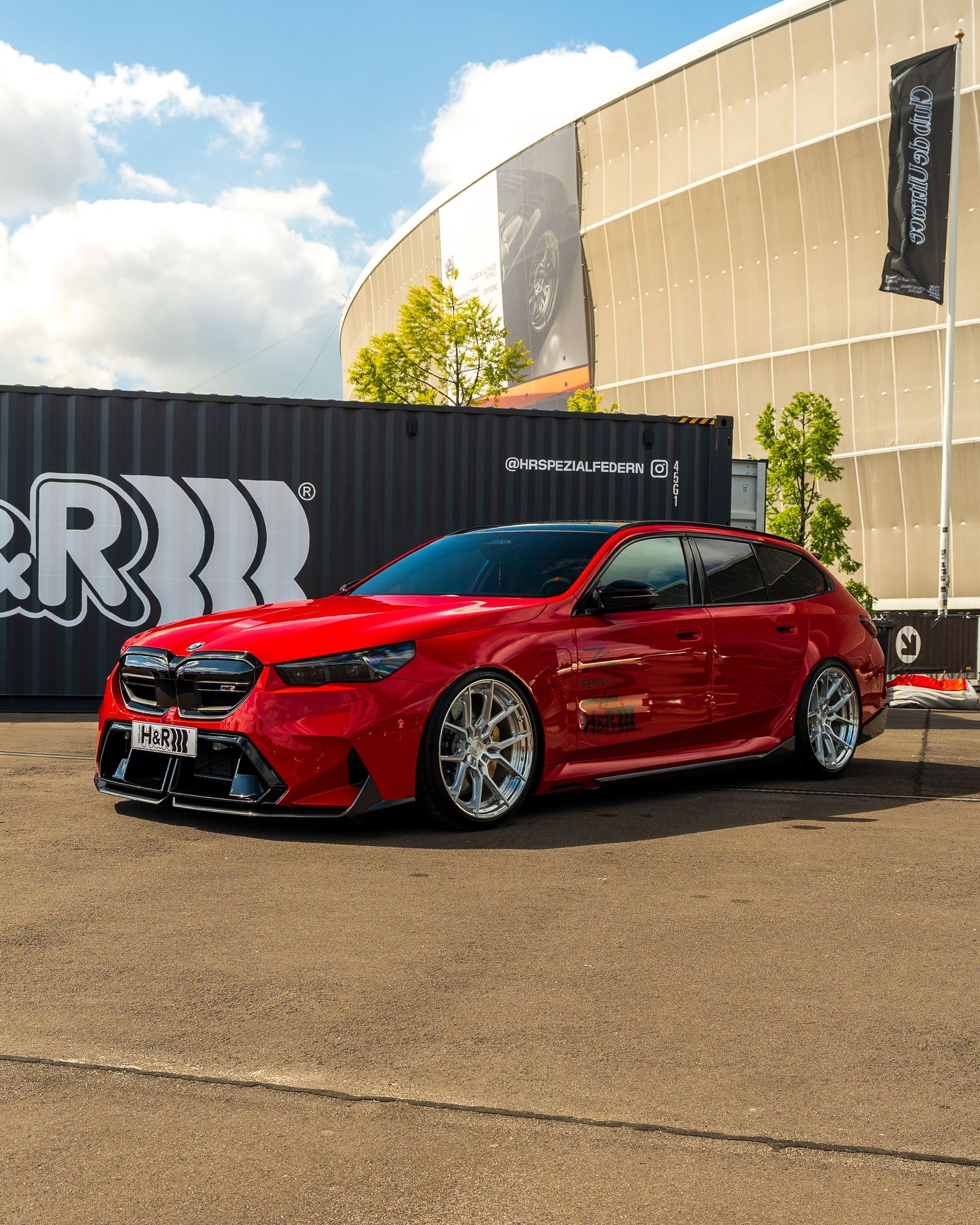 Customized red BMW M5 with lowered suspension and aftermarket wheels parked in front of an arena building.