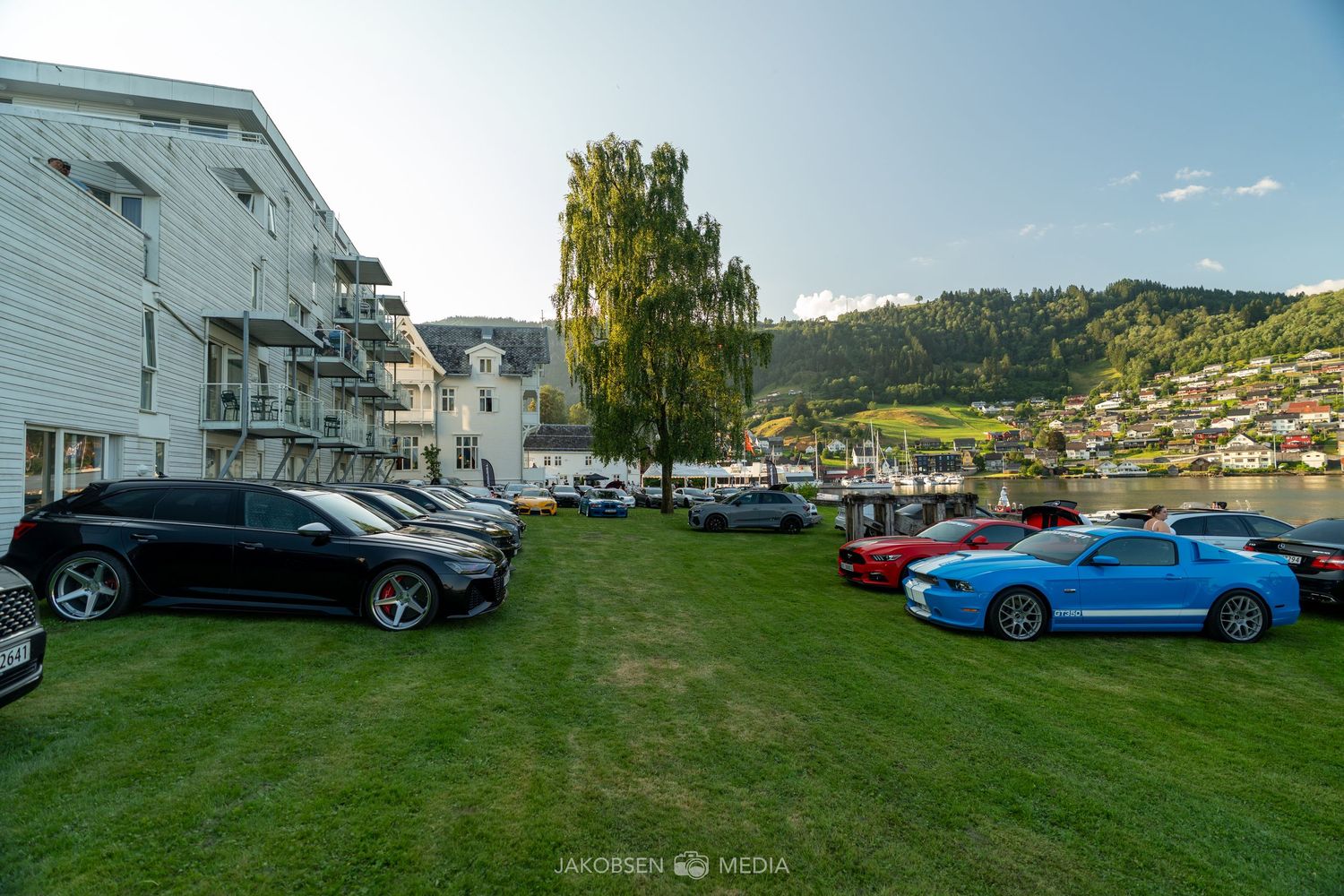 Collection of exotic cars parked on grass with mountain scenery and traditional buildings in view.