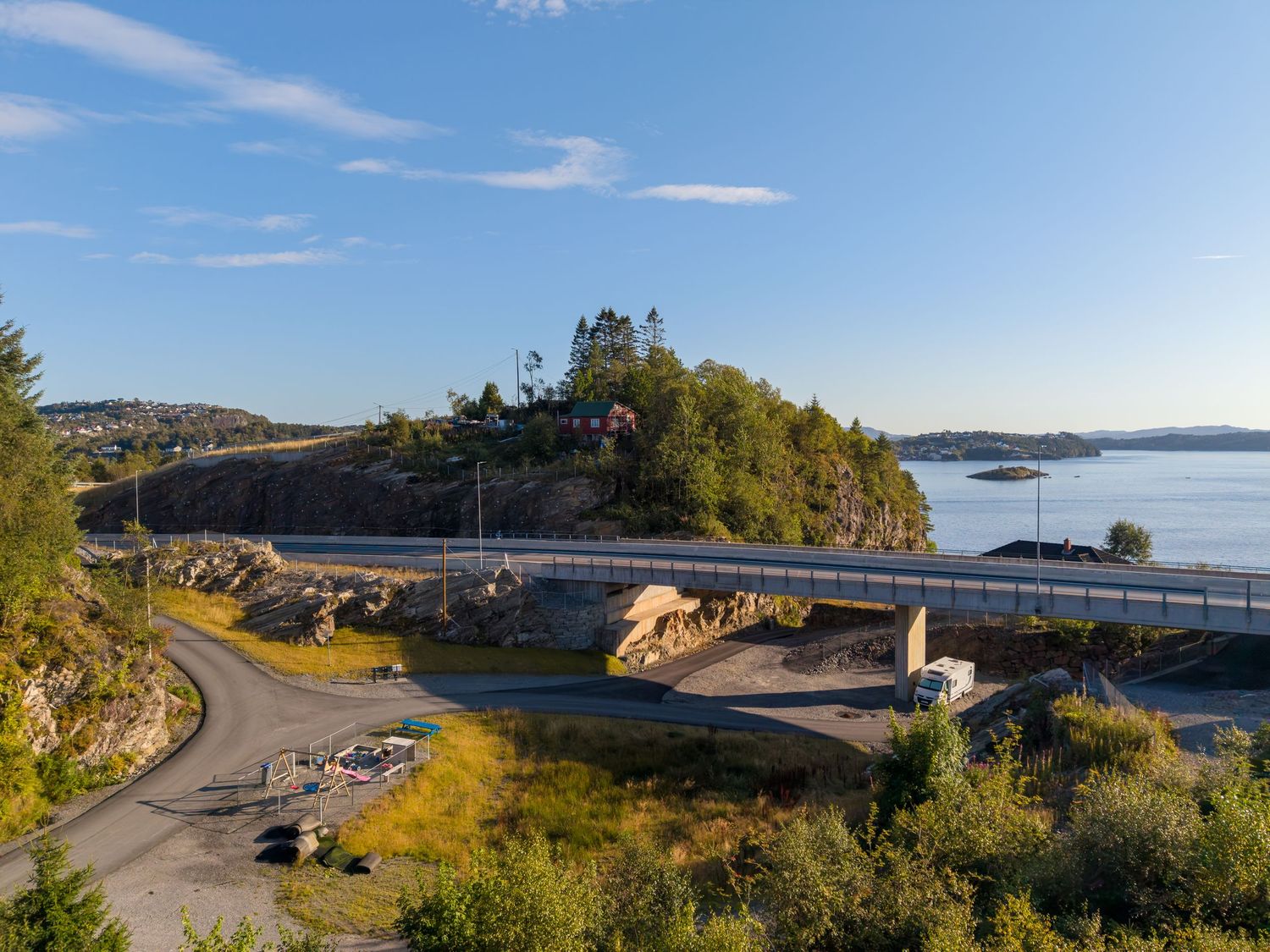 Coastal road curving past a rocky wooded hill beside a calm blue fjord on a sunny day.