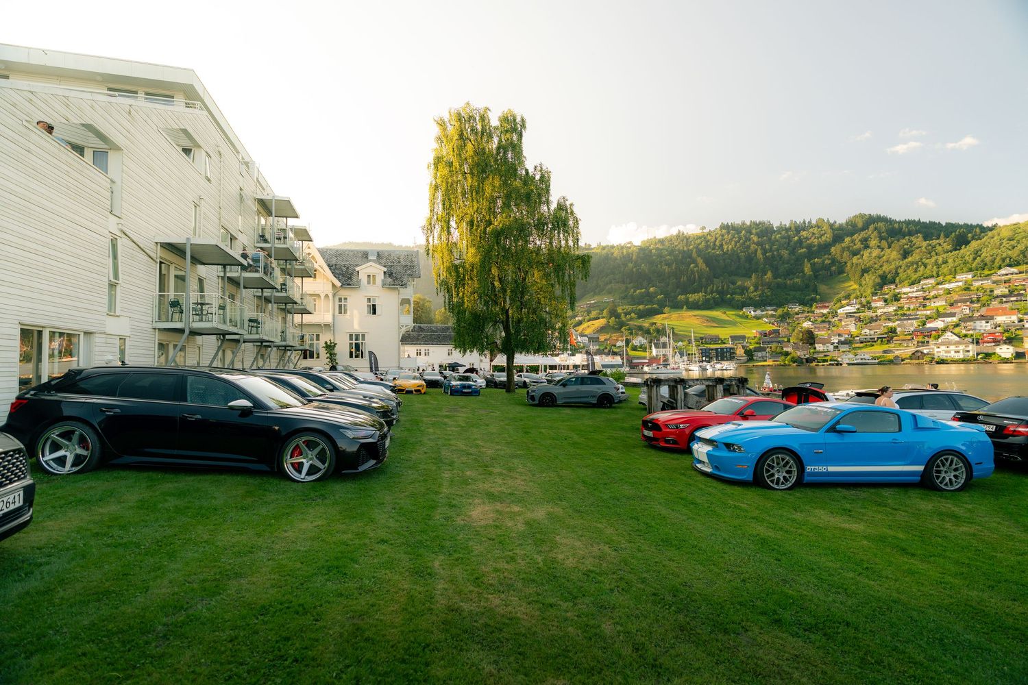 Classic and modern cars parked on a grassy lawn by a waterfront, with a blue sports coupe in front and a scenic hillside village behind.