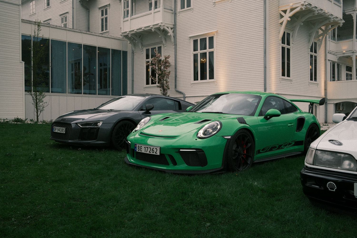 A green Porsche 911 GT3 RS sits on a lawn beside a dark gray sports car and a white car in front of a elegant white mansion.