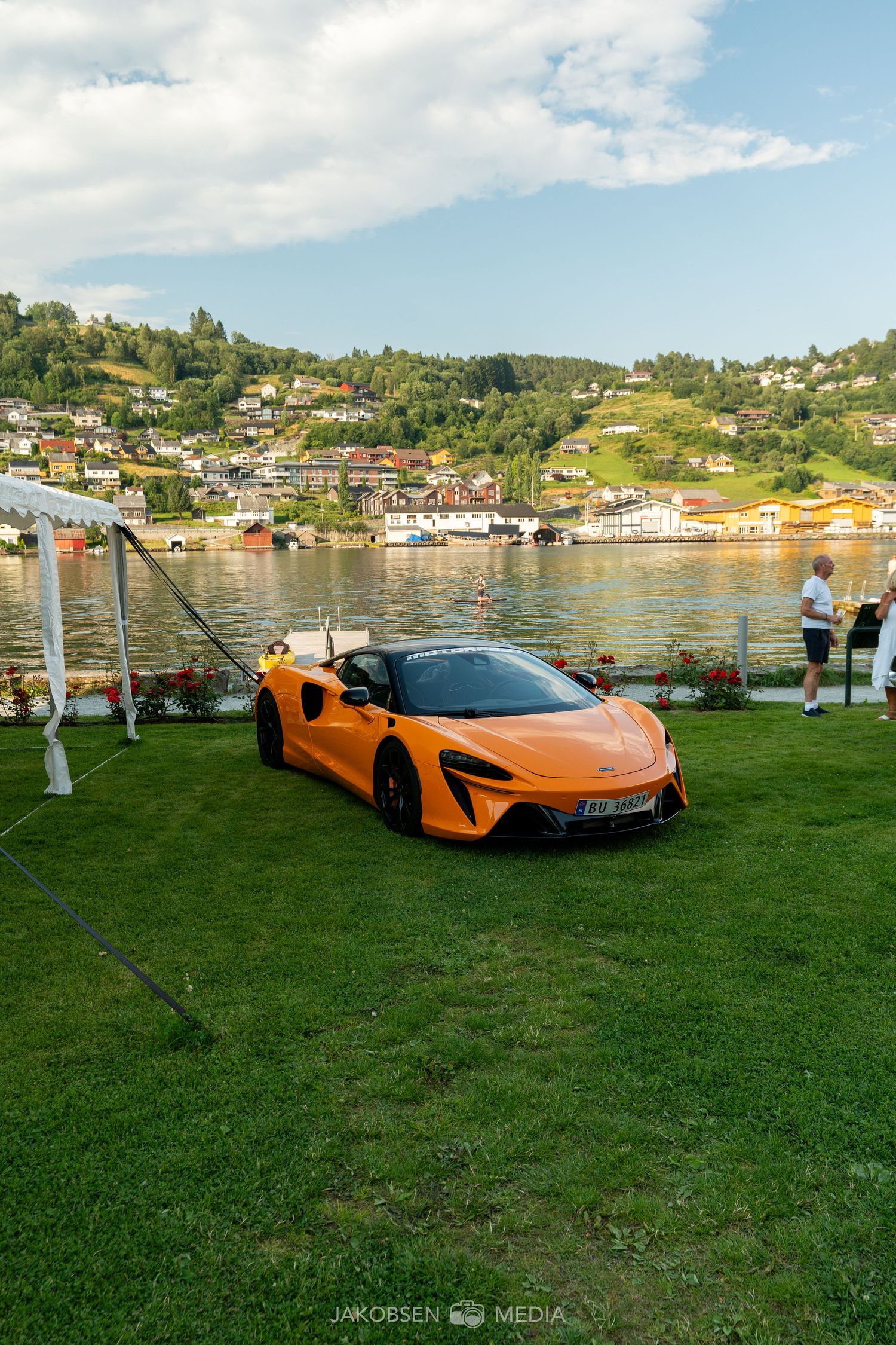 Orange McLaren sports car parked on grass with waterfront and colorful buildings in background.